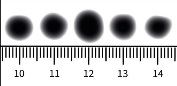 Fünf unscharfe schwarze Punkte unterschiedlicher Größe über einem Lineal mit Zentimeter-Skala (10–14 cm)